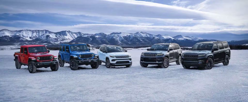A lineup of five diverse Jeep SUVs and trucks in various colors parked in a row on a vast, snow-covered plain with a majestic, snow-capped mountain range in the background.