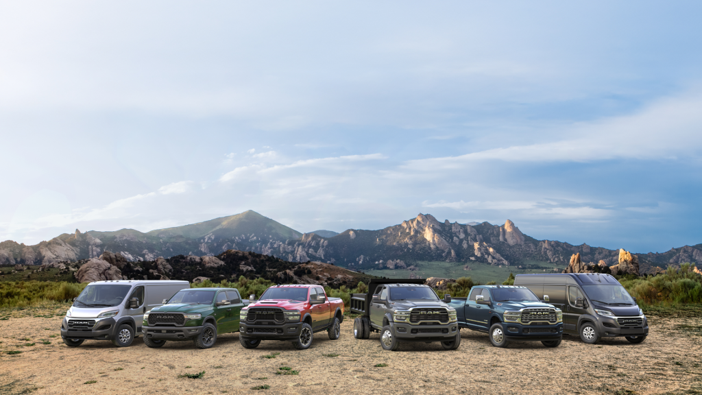 A lineup of various RAM commercial vehicles, including pickup trucks, a dump truck, and cargo vans in different colors, parked in a row on a dirt field with a scenic mountain range under a cloudy sky in the background.